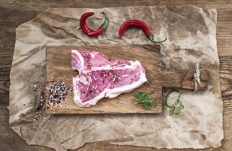 Raw meat t bone steak on serving board with red chili peppers  spices  fresh rosemary over oily craft paper and rustic wooden background
