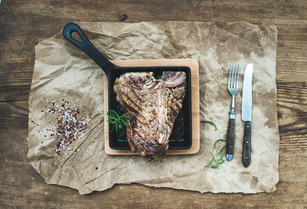 Cooked meat t bone steak in grilling pan with spices and fresh rosemary on oily craft paper over rustic wooden background