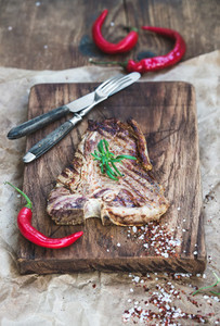 Cooked meat t bone steak on serving board with red chili peppers  spices  fresh rosemary over oily craft paper and rustic wooden background  selective focus