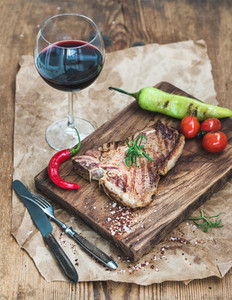 Cooked meat t bone steak on serving board with roasted tomatoes  charlestone green pepper  chili  fresh rosemary  spices and glass of red wine over rustic wooden background