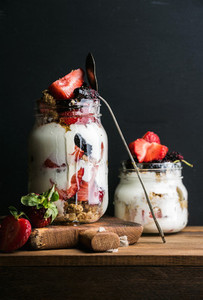 Yogurt oat granola with strawberries mulberries honey and mint leaves in tall glass jar on black backdrop