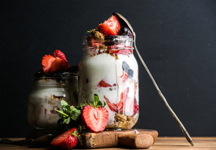 Yogurt oat granola with strawberries  mulberries  honey and mint leaves in tall glass jar on black backdrop