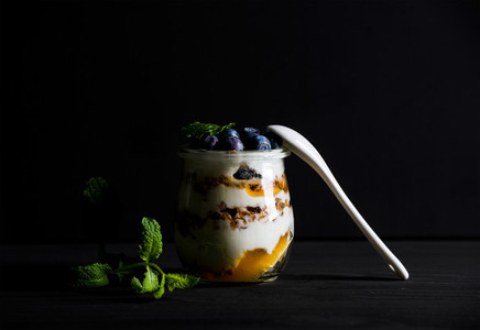 Yogurt oat granola with jam  blueberries and mint leavesin glass jar on black backdrop