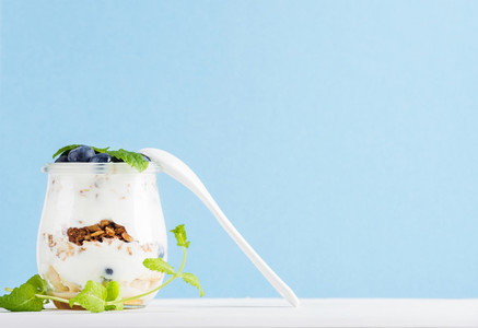 Yogurt oat granola with jam blueberries and green leaves in glass jar on pastel blue backdrop