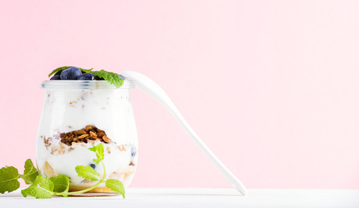 Yogurt oat granola with jam blueberries and green leaves in glass jar on pastel pink backdrop