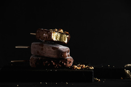 Stack of chocolate dipped popsicles with chipped nuts on dark wooden board over black background