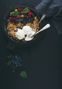Healthy breakfast Oat granola crumble with fresh berries seeds and ice cream in iron skillet pan on dark wooden board over black backdrop