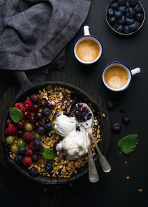Healthy breakfast  Oat granola crumble with fresh berries  seeds and ice cream in iron skillet pan on dark wooden board  coffee over black backdrop