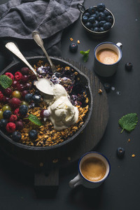 Healthy breakfast  Oat granola crumble with fresh berries  seeds and ice cream in iron skillet pan on dark wooden board  coffee over black backdrop