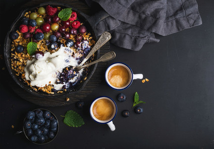 Healthy breakfast Oat granola crumble with fresh berries seeds and ice cream in iron skillet pan on dark wooden board coffee over black backdrop top view