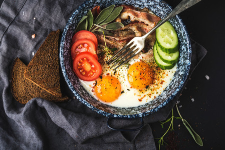 Breakfast set  Pan of fried eggs with bacon  fresh tomato  cucumber  sage and bread on dark serving board over black background