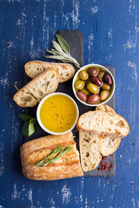 Mediterranean snacks set  Olives  oil  herbs and sliced ciabatta bread on black slate stone board over painted dark blue background