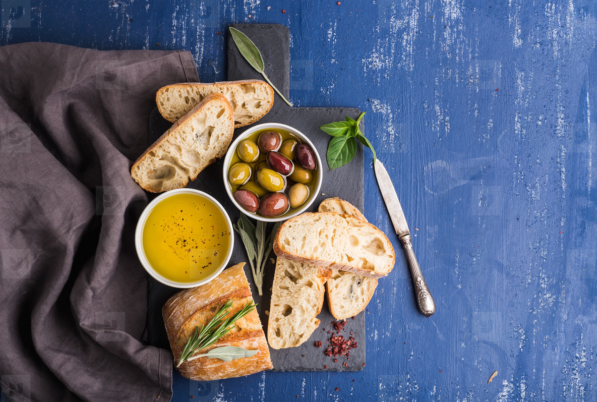 Mediterranean snacks set. Olives, oil, herbs and sliced ciabatta bread on black slate stone board over painted dark blue background