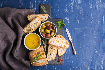 Mediterranean snacks set  Olives  oil  herbs and sliced ciabatta bread on black slate stone board over painted dark blue background