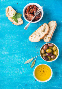 Mediterranean snacks set  Olives  oil  sun dried tomatoes  herbs and sliced ciabatta bread on over blue painted background