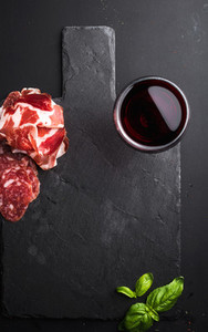 Glass of red wine  meat appetizer and basil on black  slate stone board over dark background