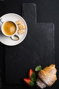 Espresso coffee cup and croissant with fresh strawberries on black  slate stone board over dark background