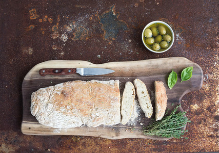 Freshly baked homemade ciabatta bread with olives basil and thyme on walnut wood board over grunge rusty metal background top view
