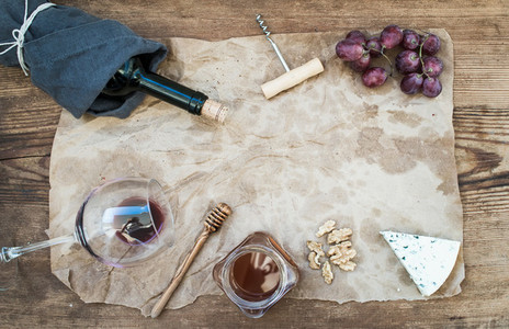 Wine and appetizer set with copy space in center  Glass of red wine  bottle  corkscrewer  blue cheese  grapes  honey  walnuts on oily craft paper over rustic wooden table  top view