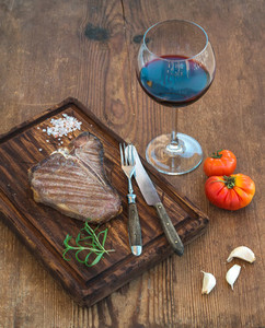Cooked meat t bone steak on serving board with garlic cloves tomatoes rosemary spices and glass of red wine over rustic wooden background