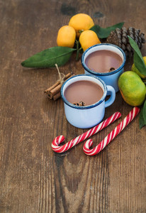 Hot chocolate in enamel metal mugs  fresh mandarins  cinnamon sticks  pine cone and candy canes over rustic wooden background
