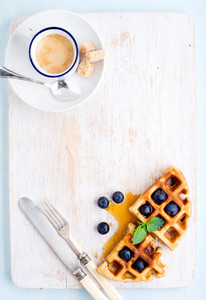 Espresso coffee cup  soft belgian waffles with fresh blueberries and marple syrup on white painted wooden board over light blue background