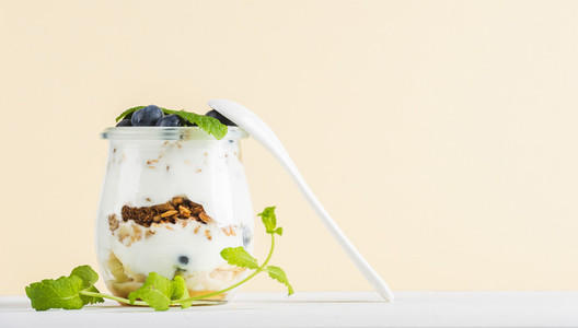 Yogurt oat granola with jam blueberries and green leaves in glass jar on pastel yellow backdrop