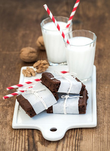 Chocolate brownie slices wrapped in paper and tired with rope glasses of milk stripe straws walnuts on white ceramic board over rustic wooden background