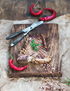 Cooked meat t bone steak on serving board with red chili peppers spices and fresh rosemary over oily craft paper rustic wooden background