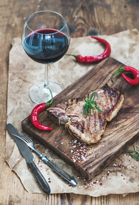 Cooked meat t bone steak on serving board with roasted tomatoes chili peppers fresh rosemary spices and glass of red wine over rustic wooden background