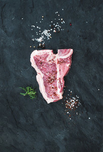 Raw fresh t bone beef steak with salt spices and rosemary over black slate stone background copy space