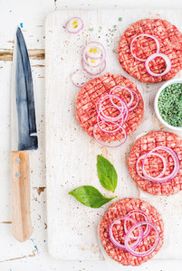 Raw ground beef meat cutlet for making burgers with onion rings spices and knife on white wooden board