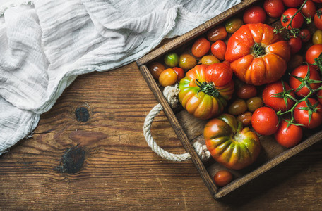 Colorful tomatoes of different sizes and kinds in dark wooden tray over rustic background copy space