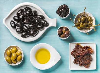 Mediterranean snack assortment  Black and green Greek olives  capers  olive oil  sun dried tomatoes over turquoise blue background