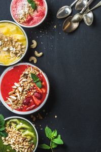 Healthy summer breakfast concept Colorful fruit smoothie bowls with nuts oat granola and mint leaves five silver spoons on black background Yellow pink red green Top view copy space