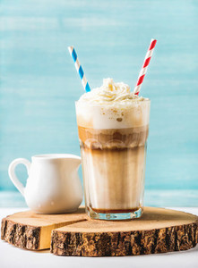 Latte macchiato with whipped cream in tall glass and pitcher on round wooden board over blue painted wall background