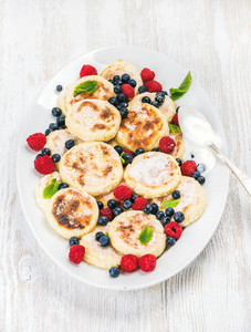 Syrniki or cottage cheese pancakes with fresh forest berries and sour cream sauce in serving dish over white wooden background
