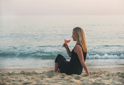 Young blond woman enjoying glass of rose wine on beach near the sea at sunset