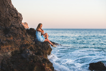 Young blond woman tourist in blue dress sittig on rocks by the sea at sunset Alanya Mediterranean region Turkey