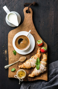 Breakfast set Two freshly baked croissants with strawberries honey cup of black coffee pitcher and spoon on rustic wooden board over dark background