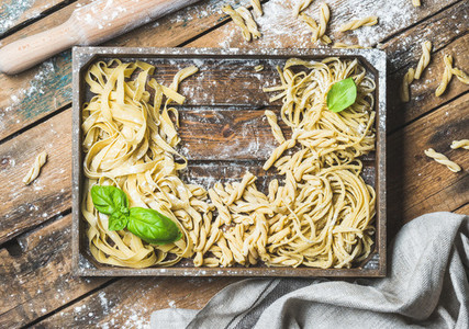 Various uncooked Italian pasta in wooden tray with basil leaves