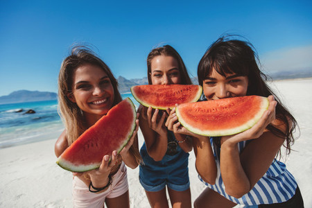 Girlfriends smiling and eating watermelon on the beach