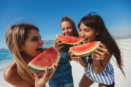 Female friends laughing and eating watermelon on the shore