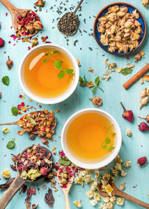 Two cups of healthy herbal tea with mint cinnamon dried rose and camomile flowers in spoons over blue background