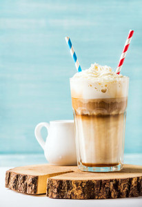 Latte macchiato with whipped cream in tall glass   two straws and pitcher on wooden board over blue painted wall background  copy space