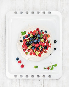 Homemade Pavlova cake with fresh garden berries on white baking tray over light wooden background
