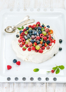 Homemade Pavlova cake with fresh garden berries and silver spoons on white baking tray over light wooden background