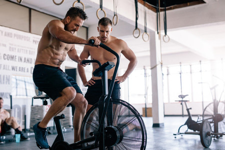 Muscular man cycling in a gym with personal trainer