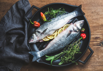 Raw uncooked seabass fish with lemon slices herbs and spices on black grilling iron pan over rustic wooden background