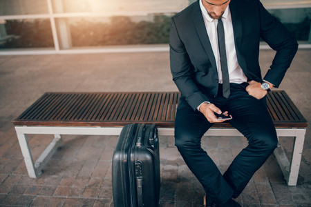 Businessman sitting on bench with suitcase and using smart phone